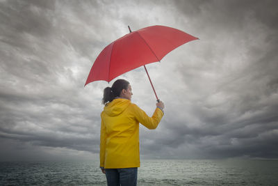 Woman looking out to see and a stormy sky, holding a red umbrella and wearing a yellow coat. 