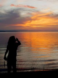 Silhouette woman photographing sea against sky during sunset