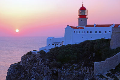 Lighthouse by sea against buildings during sunset