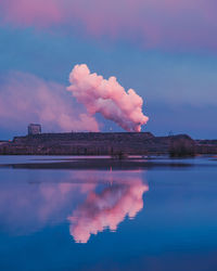 Smoke emitting from factory by lake against sky