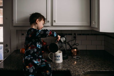 Young boy sitting on kitchen counter in pjs making coffee for dad