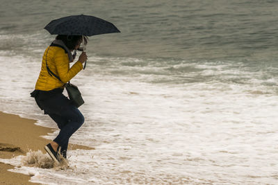 Full length of woman holding umbrella while playing with waves at beach
