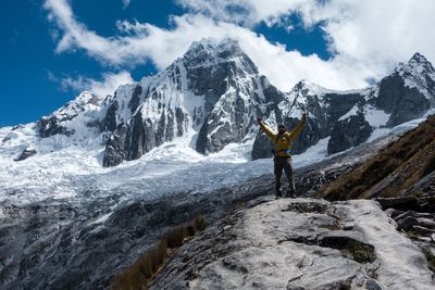 Man with arms raised standing on rock formation against snowcapped mountains