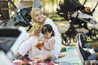 Smiling woman in hijab looking away while sitting with daughter holding teddy bear