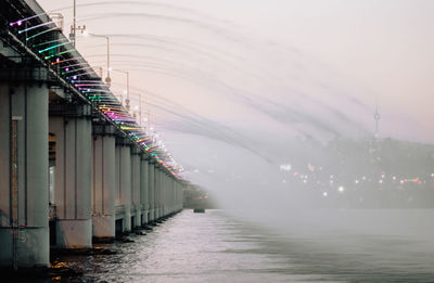 Illuminated bridge over river amidst buildings against sky