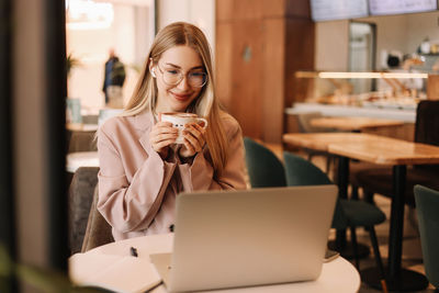 Portrait of a woman student with glasses smiling drinking coffee working online using laptop in cafe