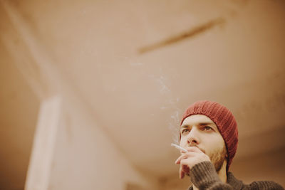 Low angle view of young man wearing knit hat smoking while standing in room