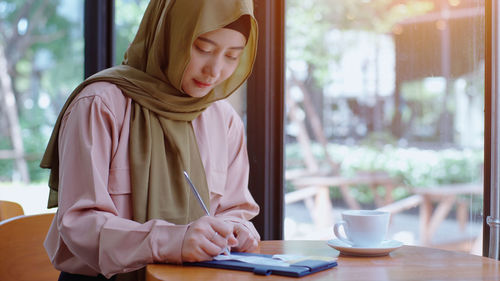 Young woman in hijab writing on paper while sitting at cafe