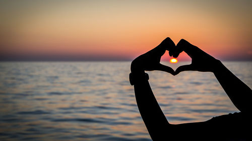 Close-up of man forming heart shape against sky during sunset