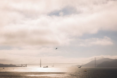 View of suspension bridge over sea against cloudy sky