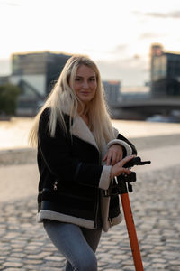 Portrait of young woman standing with push scooter on footpath in city