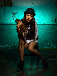 Portrait of young woman holding cleaning equipment while sitting on stool against wall