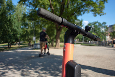 Handlebar of an electric scooter in the public park with a man riding one in the background.