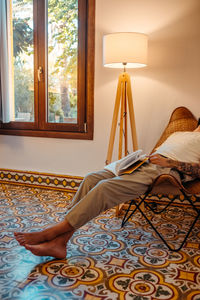 Crop unrecognizable barefooted male tourist in casual clothes relaxing on armchair with book in modern house with white walls and traditional azulejo tiled floor in mallorca