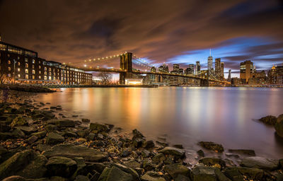 Illuminated bridge over river by buildings against sky at night