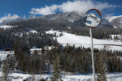 Fir trees, snow, mountains and parabolic road mirror reflecting a mountain with snow.