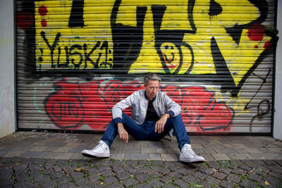 Full length portrait of young man sitting on footpath against wall