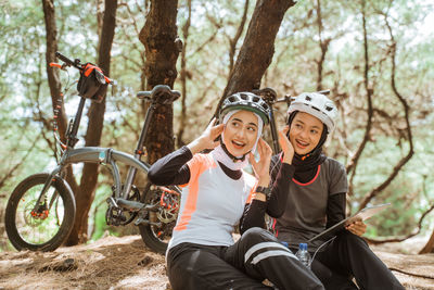 Smiling females with bicycle sitting against trees