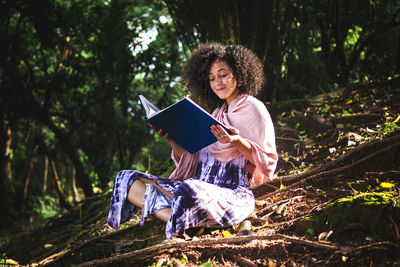 Young woman with a curly hair sitting on a floor with tree roots reading a book and smiling