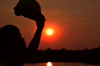 Silhouette boy holding cap against sky during sunset