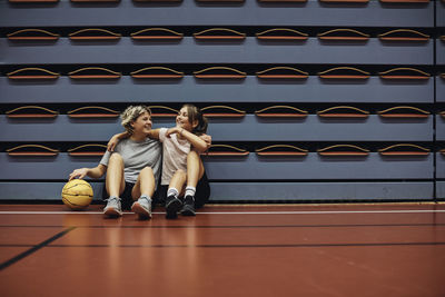 Full length of smiling girls sitting with arms around each other at basketball court in school