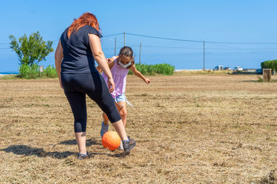Full length of mother and daughter with protective face mask playing football on field
