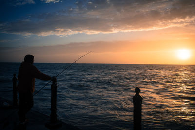 Silhouette man fishing in sea against sky during sunset