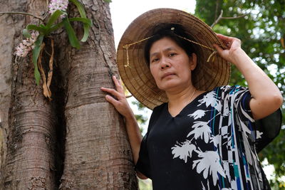 Portrait of mature woman wearing asian style conical hat by tree in forest