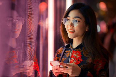 Smiling young woman using smart phone by window with reflection at night 