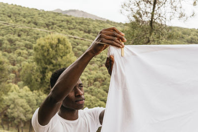 Focused african american male hanging white cotton cloth on clothesline in countryside in summer