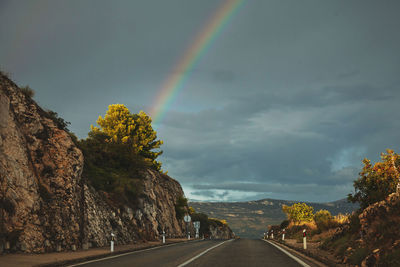 Rainbow rising in dark cloudy stormy sky over the road between mountains on sunset