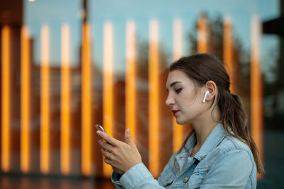 Young woman listening to music and using mobile phone while standing against glass window