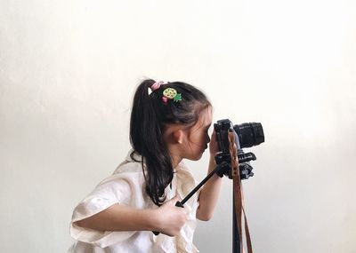 Side view of girl photographing with digital camera by white wall