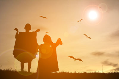 Silhouette people in halloween costume standing on field with birds flying in background during sunset