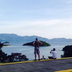 Rear view of men on roadside looking at sea and mountains against sky