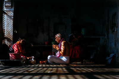 Varanasi, india - february, 2018: side view of aged indian guru performing religious ritual and praying while sitting on floor in font of male visitor in dark room with sunlight breaking through lattice window