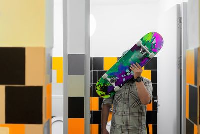 Boy covering face with skateboard in bathroom