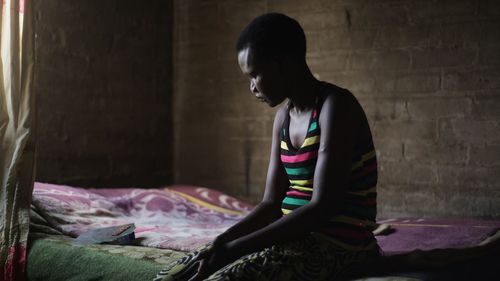 Side view of woman sitting on bed against wall at home