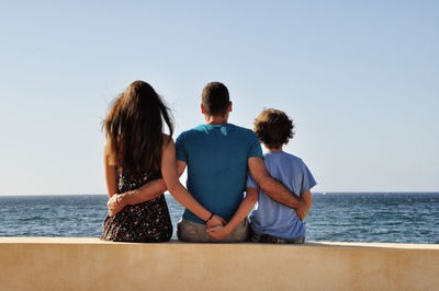 Rear view of family sitting by sea against clear sky