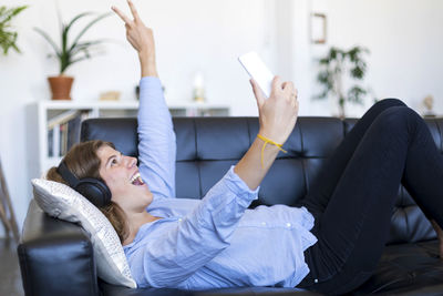 Smiling young woman using phone while lying on sofa at home