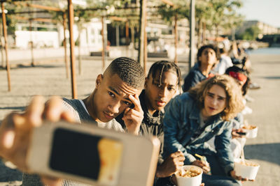 Male friends taking selfie while eating take out food on street in city