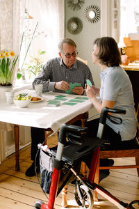 Full length of mature female healthcare worker playing cards with retired senior man at table in nursing home