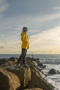Woman standing on rocks looking at the ocean, yellow coat.