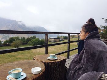 Thoughtful woman wrapped in blanket sitting by coffee cup on tree stump at mountain