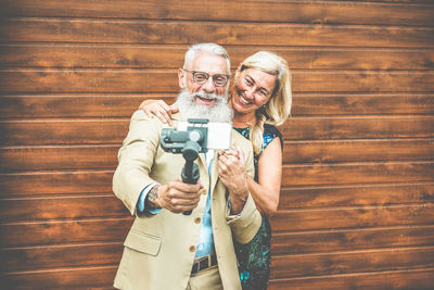 Smiling senior couple taking selfie through mobile phone while standing against wooden wall