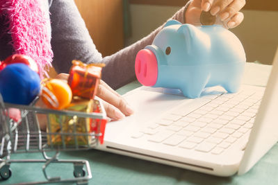 Close-up of woman inserting coin in piggy bank while using laptop