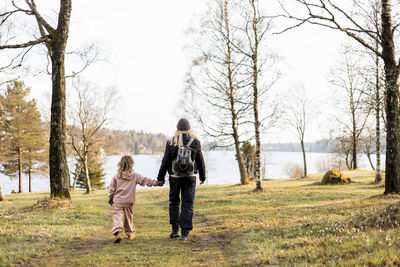 Full length rear view of mother and daughter holding hands while walking towards lake