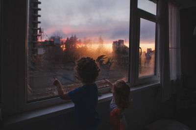 Rear view of woman looking through window at sunset