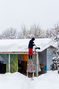 A guy shovels snow off a roof on a winter day in a snowfall