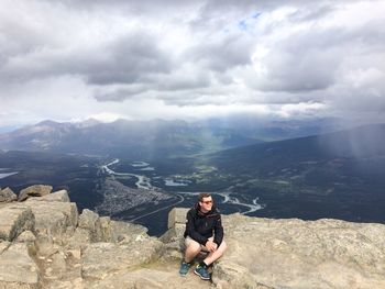 Man sitting by rocks on mountain against cloudy sky